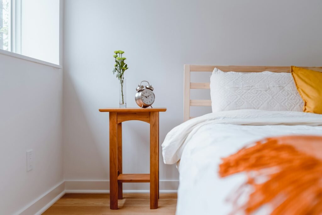 Nightstand with a plant beside a bed with white and orange linens.