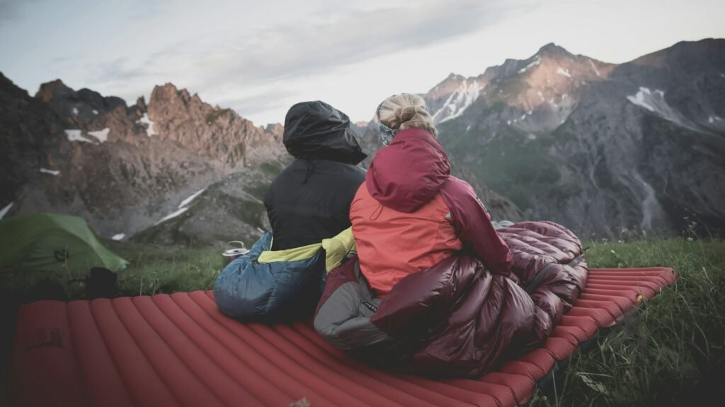 Two campers wrapped in a sleeping bag on a red pad facing mountain views.