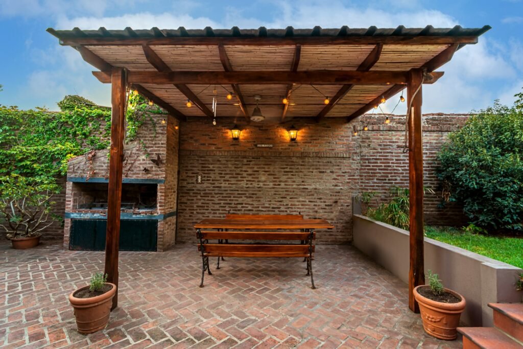 Outdoor kitchen under a wooden pergola with a bench and warm lights.