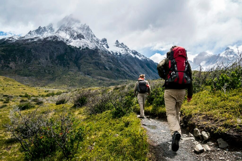Hiker with a red backpack on a trail toward snow-capped mountains.